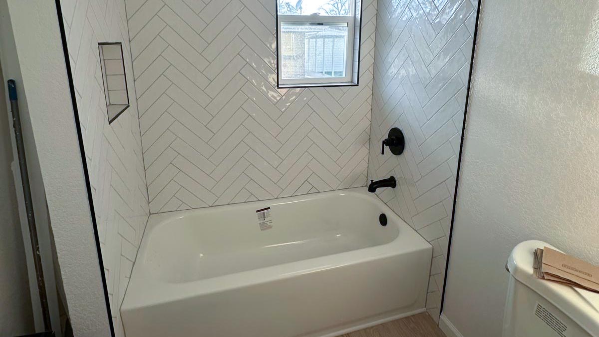 Bathtub and shower surround tiled in white herringbone with matte black fixtures and a glass shelf niche