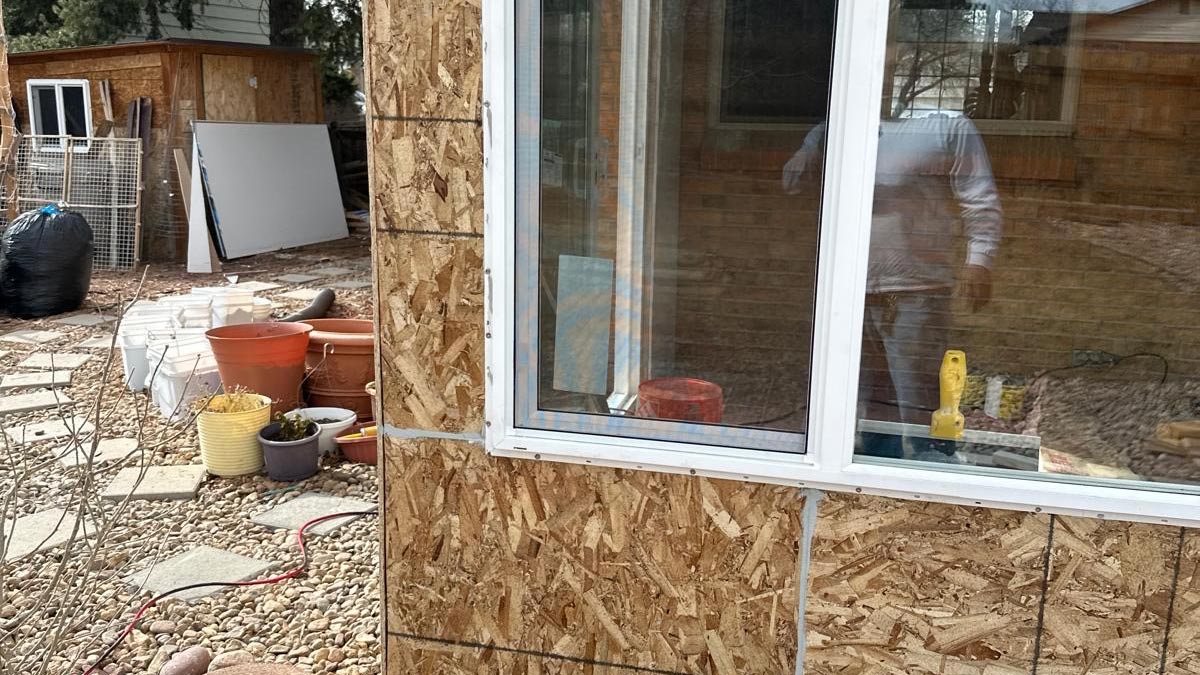 Exterior corner of a home mid-remodel showing new window openings in OSB sheathing above a brick base