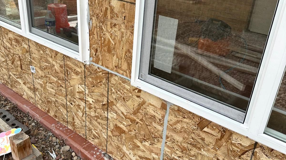 Home exterior mid-repair with siding removed, OSB sheathing exposed around vinyl windows and brick trim