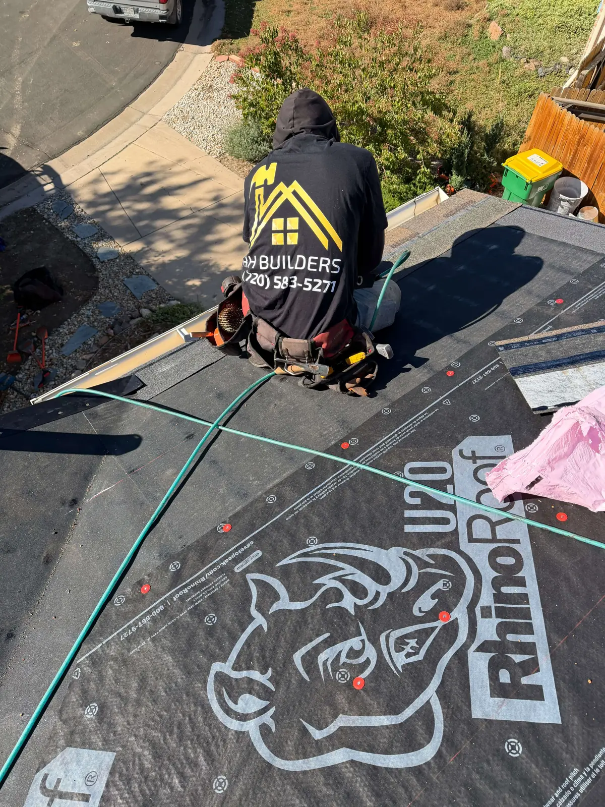 A hooded roofing worker with a tool belt kneels near the eave on printed roof underlayment.