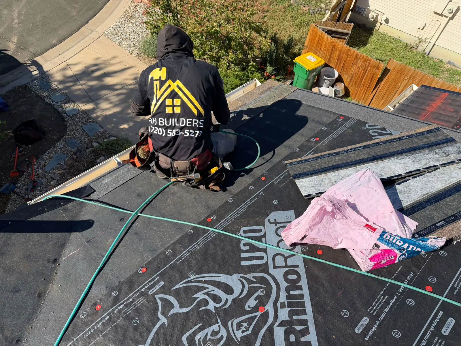 A worker kneels on black roof underlayment near the eave, with opened packaging to the right.