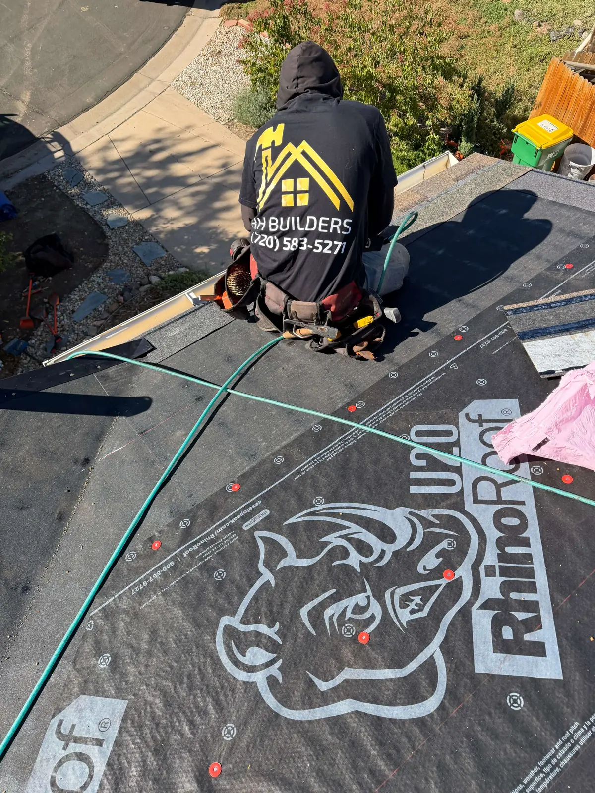 A roofing worker kneels near the roof edge with an air hose beside black printed underlayment.
