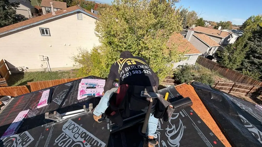 A hooded roofing worker bends over black underlayment beside wrapped shingle bundles.