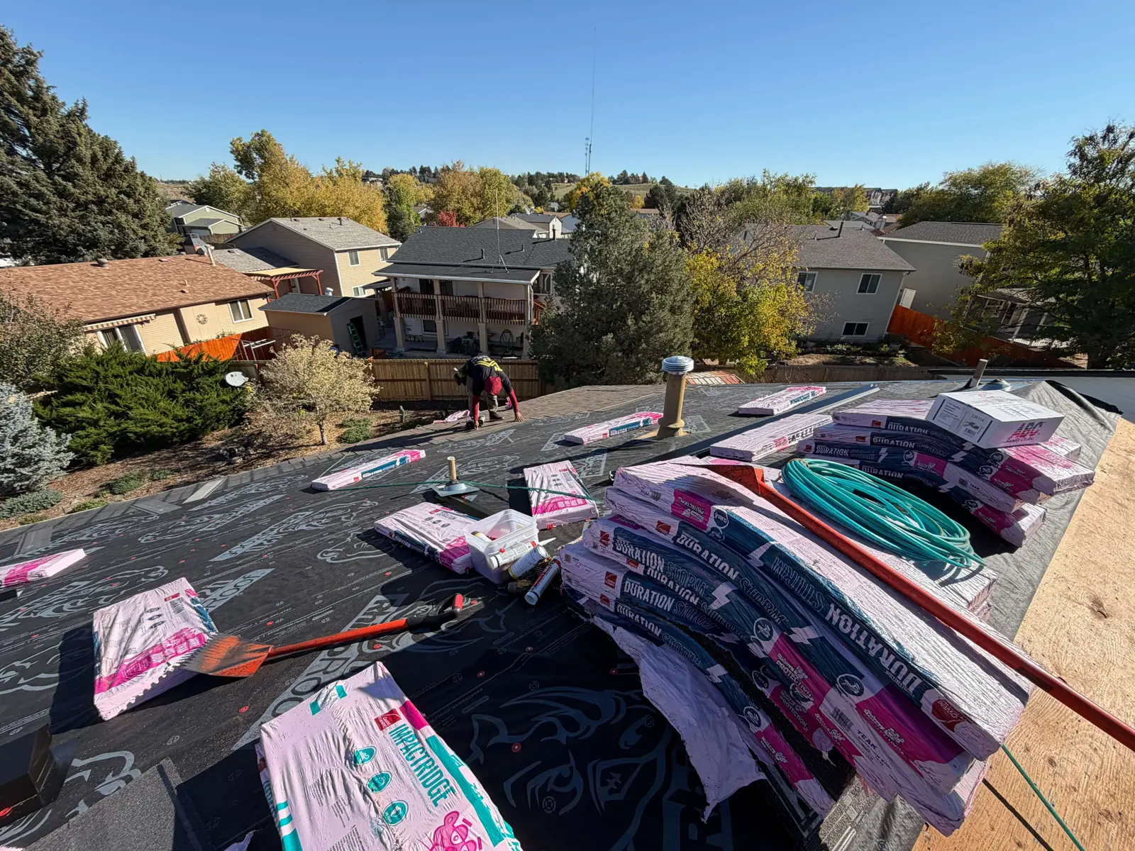Stacks of wrapped shingles on black roof underlayment, with a worker farther down the slope.