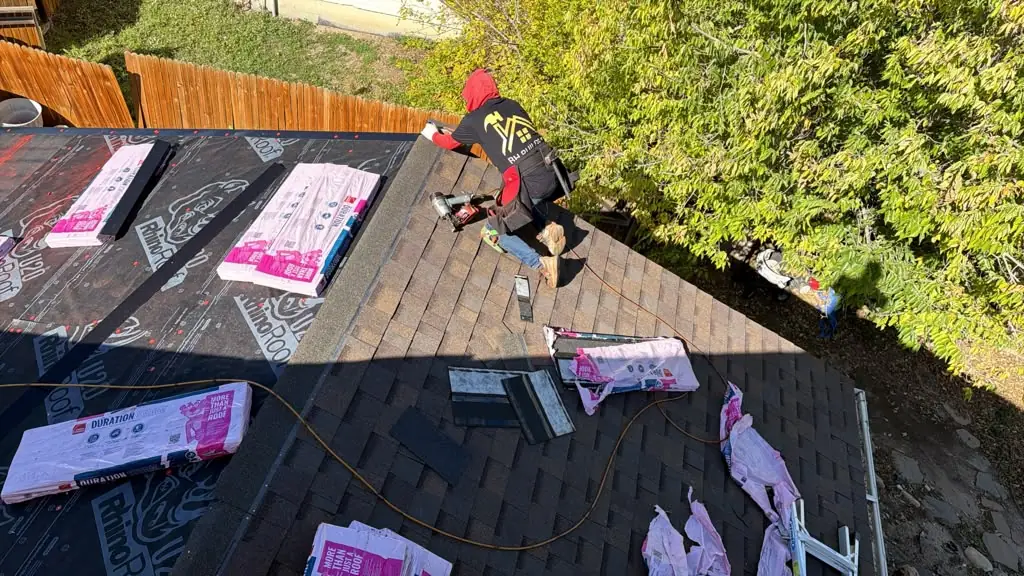 A worker in a red hood installs brown-toned shingles beside exposed black underlayment.