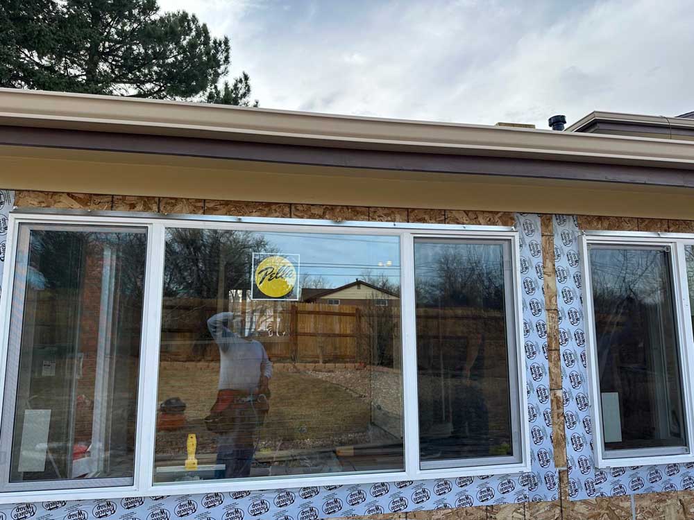 Large residential picture window installed while house wrap, flashing and roof-edge drainage remain visible