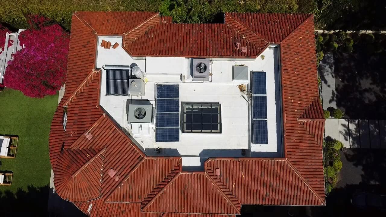 Top-down aerial of a clay tile roof surrounding a white TPO flat section with solar panels and skylight